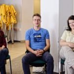 Młody mężczyzna siedzi pomiędzy dwoma młodymi, uśmiechniętymi kobietami i ze zdziwieniem spogląda w obiektyw.