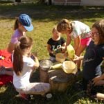 Pięć dziewczynek siedząc na trawie, przygotowuje ciasto w naczyniach leżących na pniu drzewa.