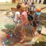 Trojka dzieci tańczy w kałuży farby boso, w tle grupa dzieci przygląda się im.