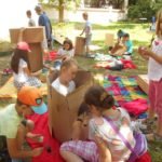 Na pierwszym planie dziewczynka siedząca w kartonie oraz trójka dziewczynek malująca ten karton. W tle grupa dzieci bawiąca się kartonami.