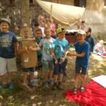 Grupa sześciu chłopców uśmiecha się wprost do obiektywu. Jeden chłopiec ubrany jest w karton. W tle grupa dziewczynek.