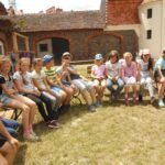 Duża grupa dzieci siedząca na ławce w półokręgu. W tle zabudowania zamkowe