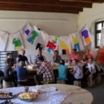 Duża grupa dzieci, młodzieży i dorosłych siedząca przy dużej drewnianej ławie. W tle za nimi rozwieszone na ścianie kolorowe plakaty.