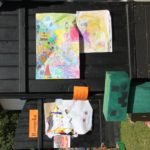 Czarna, lśniąca drewniana ława, na której leżą kolorowe obrazki malowane kredkami przez dzieci.