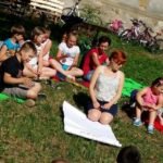 Duża grupa dzieci siedząca na trawie wokół młodej kobiety z dużym arkuszem papieru.