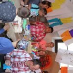 Grupa dorosłych i dzieci siedząca przy stole. Nad nimi wiszą arkusze papieru z rysunkami ubrań.