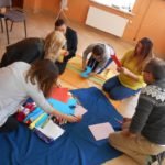 Pięć kobiet i mężczyzna, siedząc na podłodze, wycinają ozdoby z kolorowego papieru.