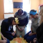 Cztery kobiety i mężczyzna w koronach na głowie z papieru kolorowego klęczą w kręgu na kocu.