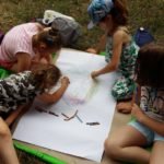 Cztery małe dziewczynki rysują kredkami na dużym arkuszu papieru, siedząc na kocu.