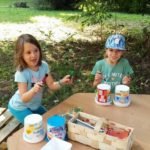 Dwie małe dziewczynki trzymające w rękach mazaki siedzą na ławce przy stoliku, na którym stoi koszyk po truskawkach i plastikowe kubki.