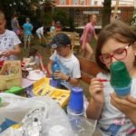 Na pierwszym planie dziewczynka i dwóch chłopców ozdabiający plastikowe kubki. W tle duża grupa dzieci.