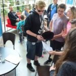 Grupa młodzieży stoi w holu muzeum i spogląda na kartkę papieru. Na bocznym planie widać stół z kartami do gry.