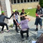 Grupa młodzieży wygłupia się z balonami w ręku na brukowanym placu. Obok nich na ławce siedzi kobieta w sukni balowej.