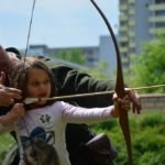 Kilkuletnia dziewczynka z pomocą instruktora napina cięciwę łuku, przygotowując się do strzału.