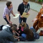 Na pierwszym planie trzy dziewczyny i chłopak siedzą na schodach plecami do obiektywu, przed nimi stoi dziewczyna oraz dwóch chłopaków i rozmawiają ze soba.