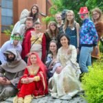 Grupa młodzieży, z której część jest ubrana w szaty z minionych epok siedzi na chodniku obok dużego soczyście zielonego krzewu.