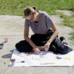 Kobieta siedząca na betonie przy arkuszu papieru nakleja na niego wycięte elementy z kolorowych gazet